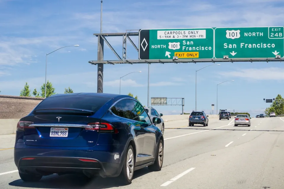 Dark blue Tesla sedan driving on freeway with green signs for San Francisco.